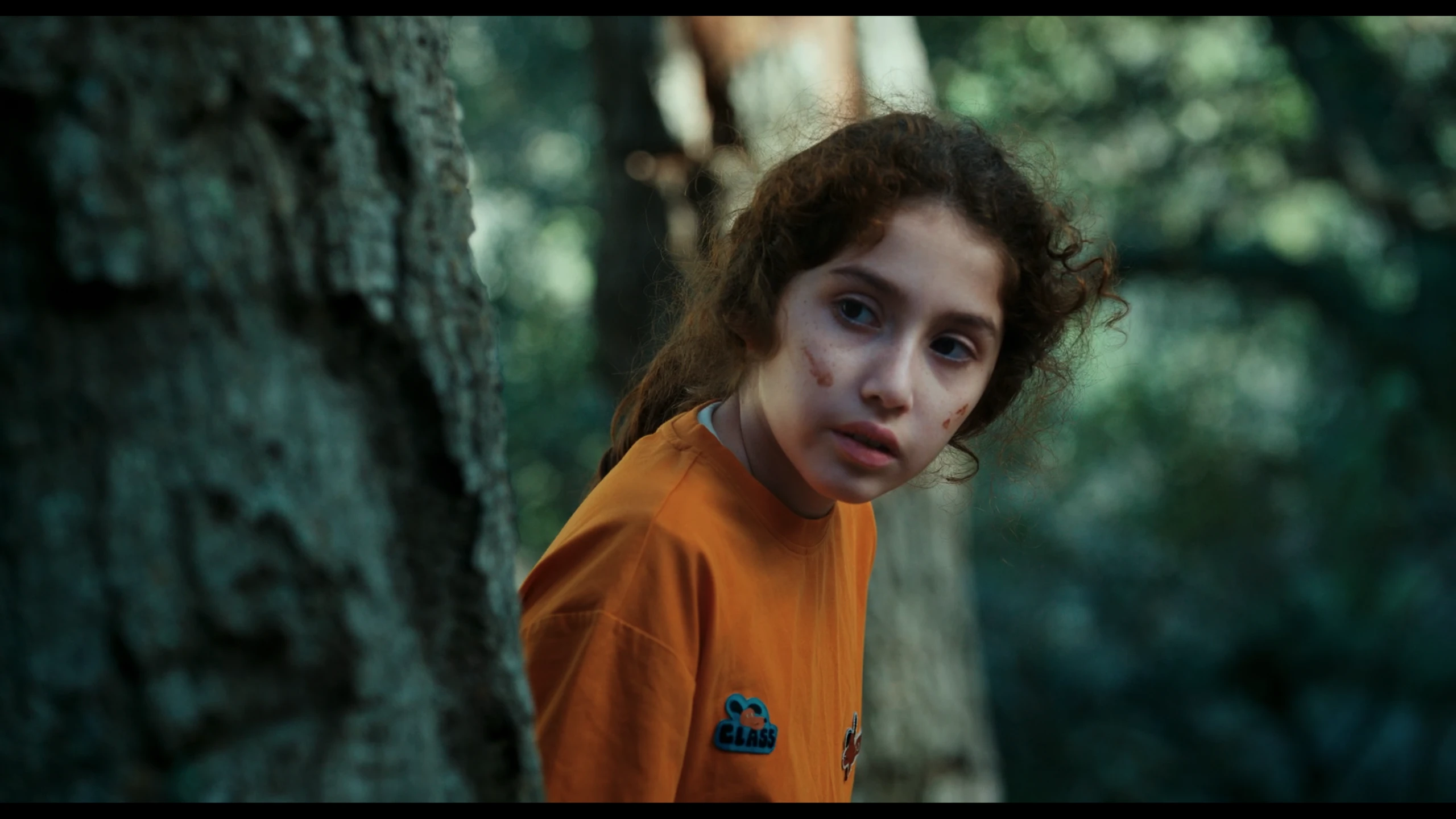 The girl in orange looks back from behind a tree in Don’t Peek