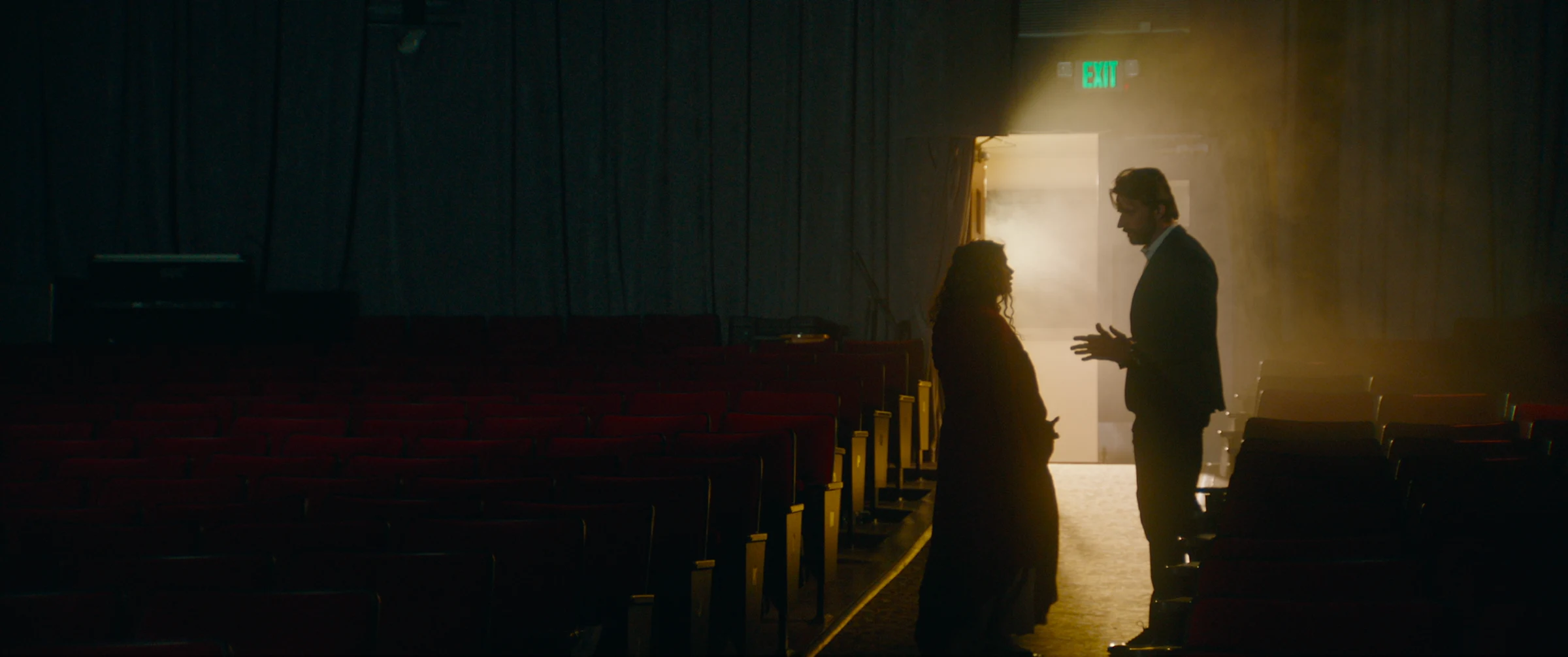Two silhouetted performers meet in a theatre aisle flooded with warm backlight in Witch Lament